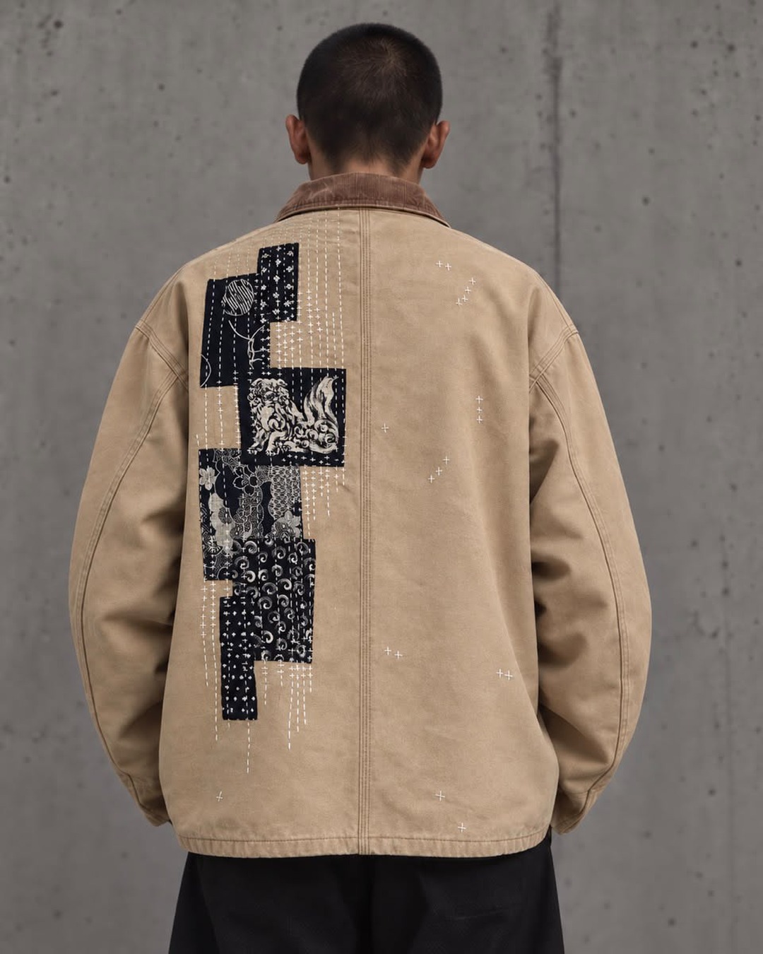 Back view of a tan work jacket with black textile patches arranged vertically and secured with white visible running stitches.